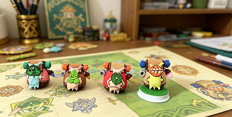 May include: Four colorful, whimsical figurines resembling backpacks with faces, set on a game board. Each figure has unique features, including leaves, orange accents, and different colored bodies. The background includes art supplies and a wooden shelf.