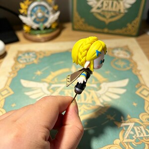 May include: A small figurine of a character with blonde hair and a blue headband, held by a hand. The figure has a brown cape and is attached to a thin wire. In the background, a box and a decorative item with the word "Zelda" are visible.