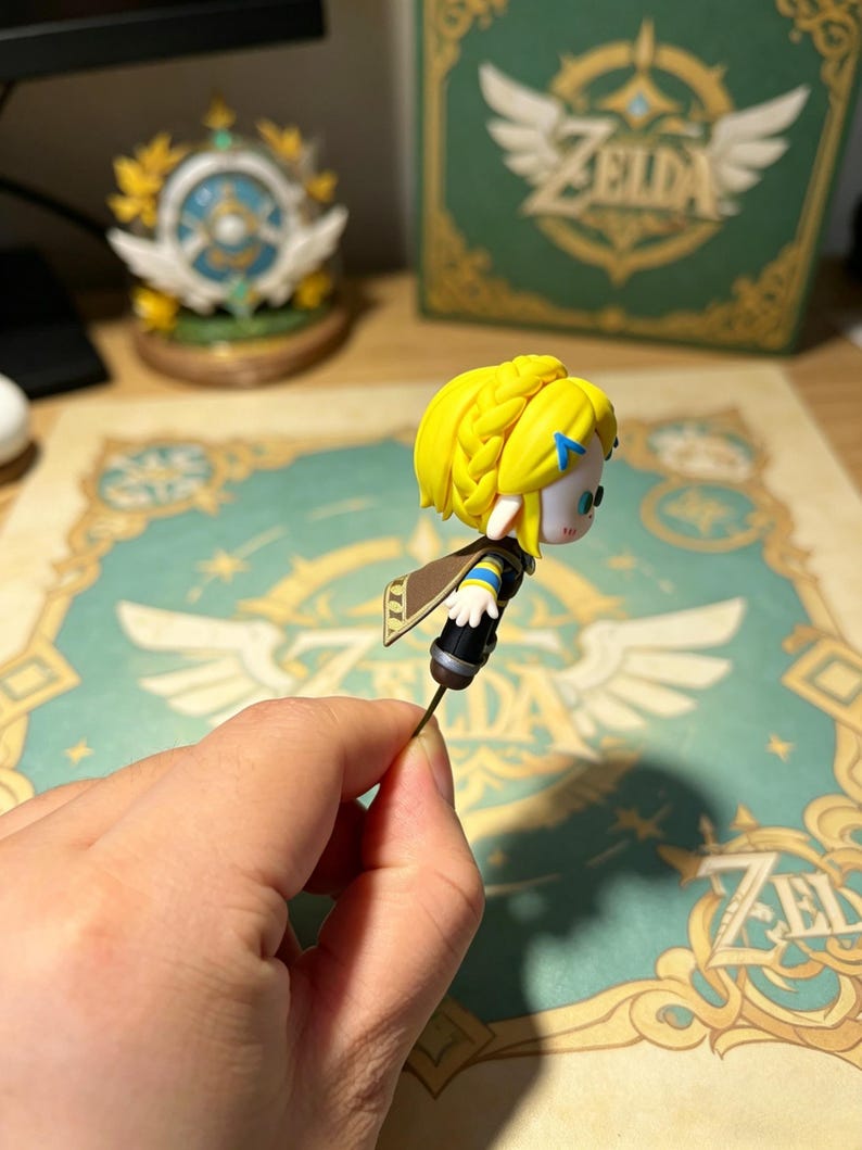 May include: A small figurine of a character with blonde hair and a blue headband, held by a hand. The figure has a brown cape and is attached to a thin wire. In the background, a box and a decorative item with the word "Zelda" are visible.