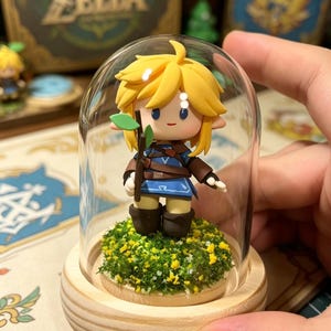 May include: A miniature figurine of a character with blonde hair, wearing a blue tunic and brown boots, holding a small green leaf, encased in a glass dome. The figurine stands on a bed of green and yellow faux grass, set on a wooden base. The word "ZELDA" is visible in the background.