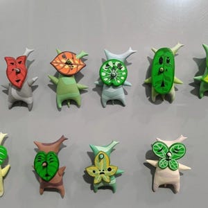 May include: Ten handmade Korok figures are displayed, each with a distinctive leaf-shaped mask and body. The figures are in various colours, including green, red, and yellow, and are set against a grey background.