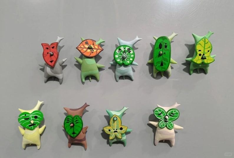 May include: Ten handmade Korok figures are displayed, each with a distinctive leaf-shaped mask and body. The figures are in various colours, including green, red, and yellow, and are set against a grey background.