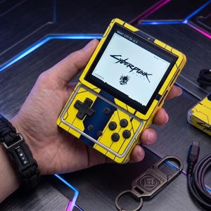 May include: A handheld gaming console with a bright yellow and blue casing featuring Cyberpunk branding on the screen. The device includes a black directional pad, four action buttons, and a small display showing the text Night City OS v1.3.