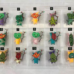 May include: A clear plastic organiser containing fifteen colourful, handmade paper figures. Each figure has a unique leaf-shaped face and holds a small branch with red berries. The figures are in various colours, including green, yellow, and blue.