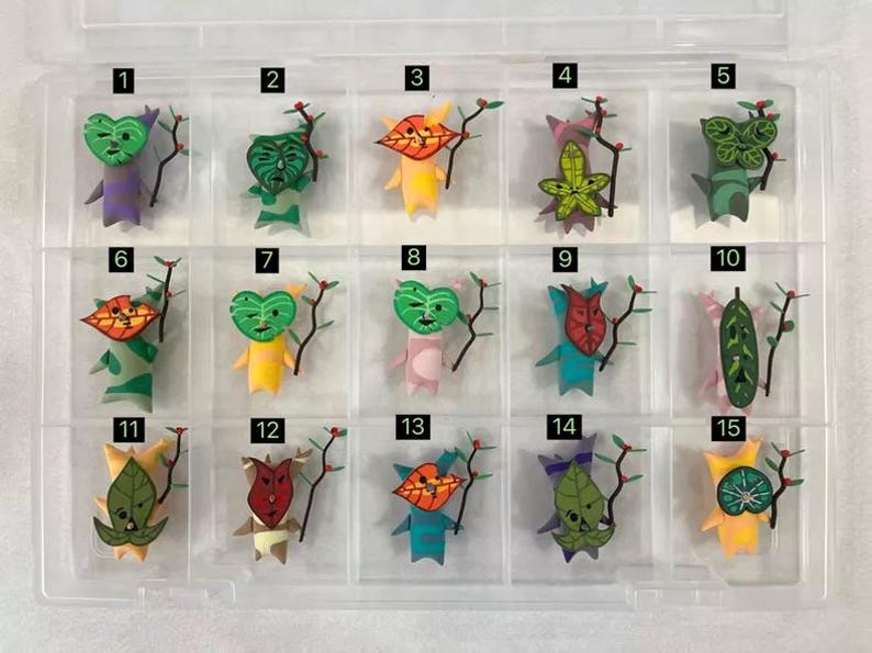 May include: A clear plastic organiser containing fifteen colourful, handmade paper figures. Each figure has a unique leaf-shaped face and holds a small branch with red berries. The figures are in various colours, including green, yellow, and blue.