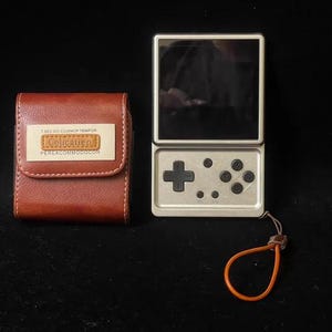 Pode incluir: Uma consola de jogos portátil prateada com ecrã preto, um pad direcional e quatro botões circulares, acompanhada por uma bolsa de transporte em pele castanha com uma etiqueta retangular com o texto Colication e outro texto impresso.