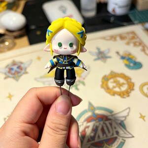 May include: A small figurine of a character with yellow hair, blue accents, and a black and blue outfit. The figurine has a light skin tone and green eyes. It is held by a person's hand.