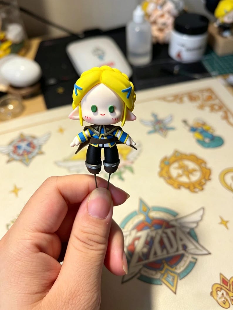 May include: A small figurine of a character with yellow hair, blue accents, and a black and blue outfit. The figurine has a light skin tone and green eyes. It is held by a person's hand.