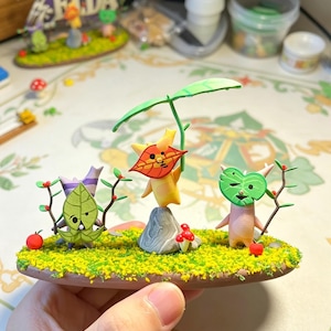 May include: A miniature diorama featuring three whimsical figures with leaf-like faces and bodies, set on a grassy base with red berries and mushrooms. The figures are in various poses, with one holding a branch. The diorama is held in a hand.