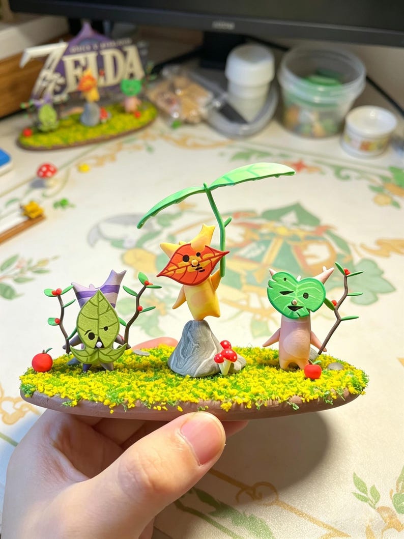 May include: A miniature diorama featuring three whimsical figures with leaf-like faces and bodies, set on a grassy base with red berries and mushrooms. The figures are in various poses, with one holding a branch. The diorama is held in a hand.