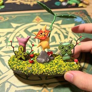 May include: A handcrafted diorama featuring three whimsical figures amidst a miniature landscape. The figures are yellow, green, and pink, with leaf-like details. The base is covered in yellow flowers and green grass, with small red berries and mushrooms.