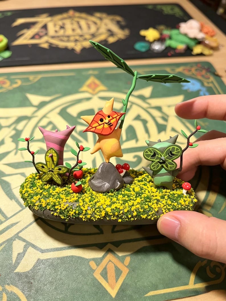 May include: A handcrafted diorama featuring three whimsical figures amidst a miniature landscape. The figures are yellow, green, and pink, with leaf-like details. The base is covered in yellow flowers and green grass, with small red berries and mushrooms.