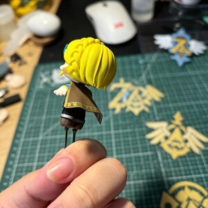 May include: A miniature figurine with a yellow hairstyle, brown cape, and black and gold accents. The figurine is held in a hand, with other crafting supplies and decorative emblems visible in the background. The figurine is approximately 2 inches tall.