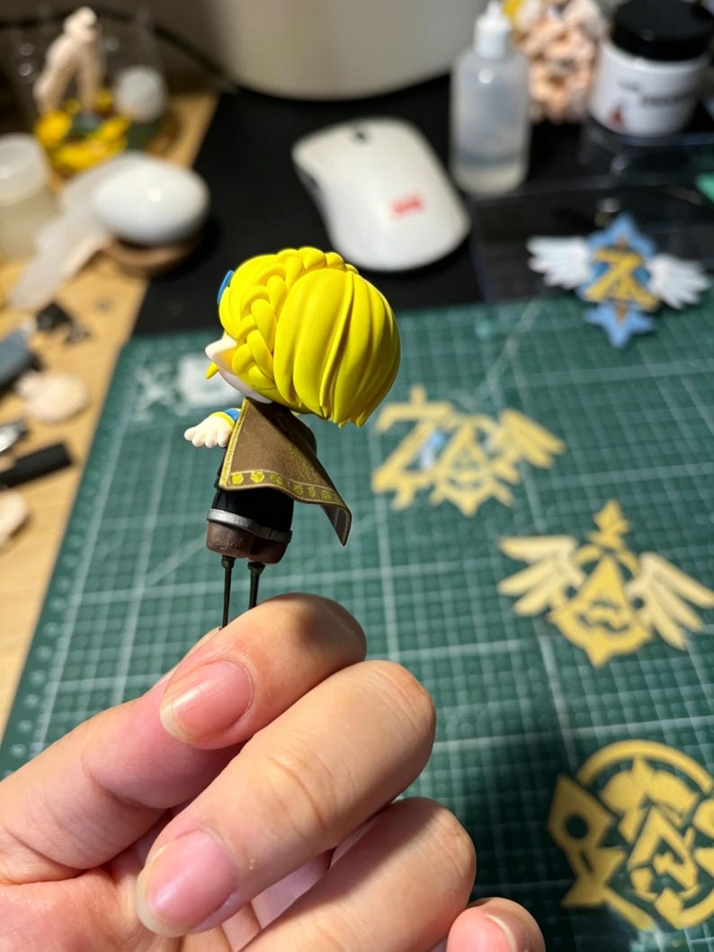 May include: A miniature figurine with a yellow hairstyle, brown cape, and black and gold accents. The figurine is held in a hand, with other crafting supplies and decorative emblems visible in the background. The figurine is approximately 2 inches tall.