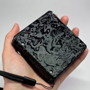 May include: A square-shaped electronic device featuring a black and grey marbled pattern on its top surface. The device has a black frame with visible silver screws at the corners and includes a black woven wrist strap attached to one side.