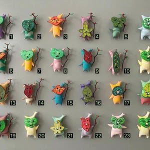 May include: A collection of twenty-four handmade figurines with leaf-like heads and small bodies. Each figure has a unique design and colour. They are arranged on a light grey surface, with each figure numbered for identification.