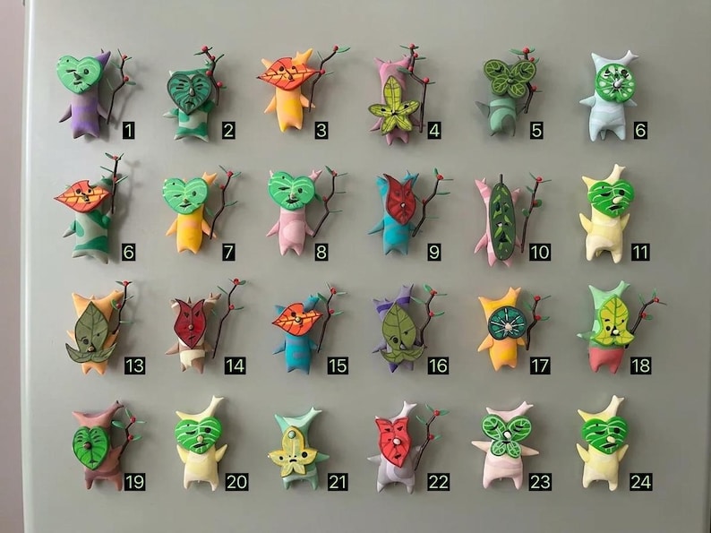 May include: A collection of twenty-four handmade figurines with leaf-like heads and small bodies. Each figure has a unique design and colour. They are arranged on a light grey surface, with each figure numbered for identification.
