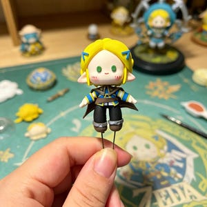 May include: A handmade figurine of a character with yellow hair, blue accents, and a brown cape. The figure is held by a person's hand. Other similar figurines are visible in the background, along with crafting tools and a wooden shelf.