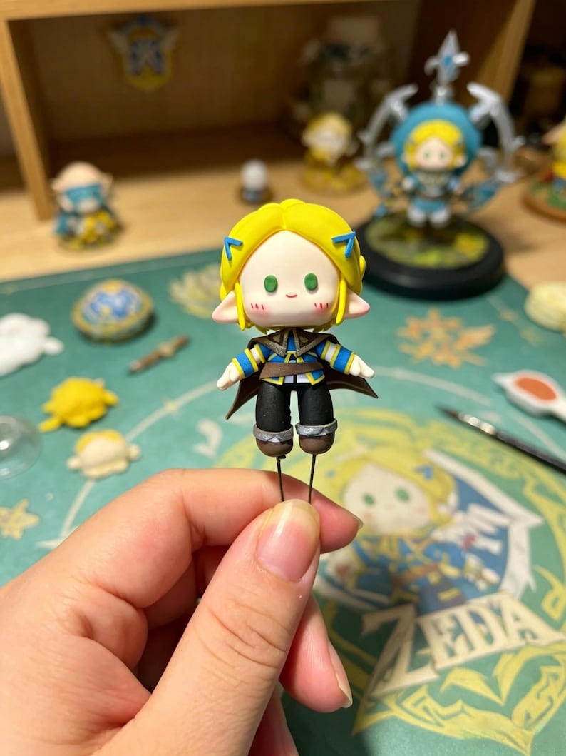 May include: A handmade figurine of a character with yellow hair, blue accents, and a brown cape. The figure is held by a person's hand. Other similar figurines are visible in the background, along with crafting tools and a wooden shelf.