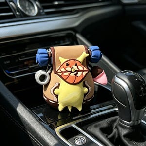 May include: A brown and yellow cartoon character car accessory. The accessory has a backpack design with blue straps and a smiling face. The accessory is sitting on a car's dashboard.
