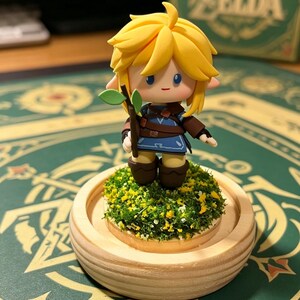 May include: A figurine of a character with yellow hair, wearing a blue tunic and brown boots, holding a small branch with leaves. The figure stands on a bed of green moss and yellow flowers, set in a wooden base. The background includes a box with the word "Zelda".