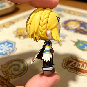 May include: A small figurine of a character with blonde hair in a braid, wearing a blue and yellow outfit with a brown cape. The figurine is held between two fingers.