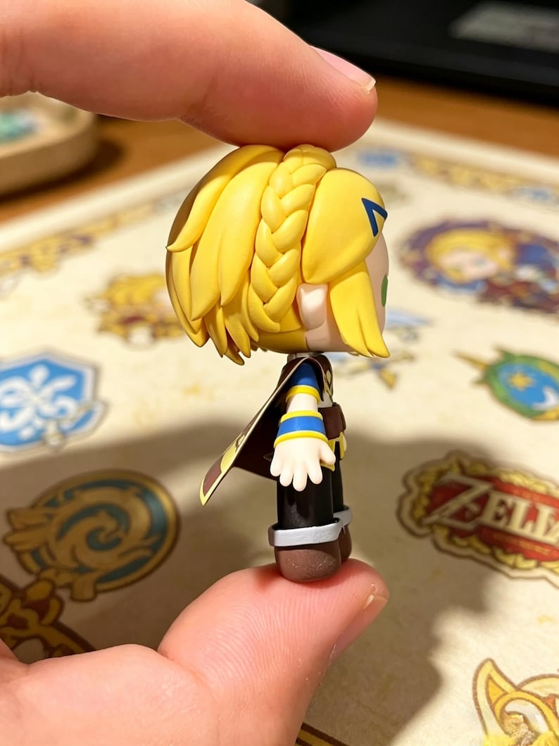 May include: A small figurine of a character with blonde hair in a braid, wearing a blue and yellow outfit with a brown cape. The figurine is held between two fingers.
