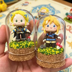 May include: Two miniature figurines of a blonde-haired character in glass domes. The figurines are dressed in blue and black outfits, standing on a bed of yellow flowers and cork bases. Small red mushroom accents are also present. The figurines are from a video game.