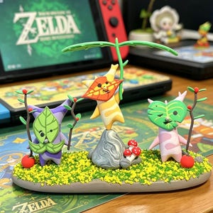 May include: A colorful figurine set featuring three Koroks from The Legend of Zelda. The figures are on a base with yellow and green detailing, with red mushrooms and apples. The background includes a Nintendo Switch with the Zelda logo.