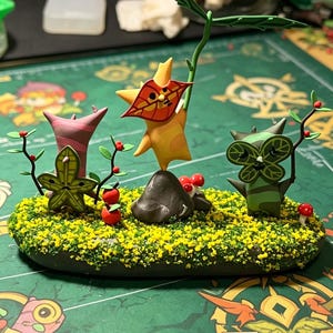 May include: A handcrafted diorama featuring three whimsical, stylized creatures amidst a miniature landscape. The scene includes a rock, small red mushrooms, and a base covered in yellow and green textured material. The creatures are in shades of green, orange, and pink.