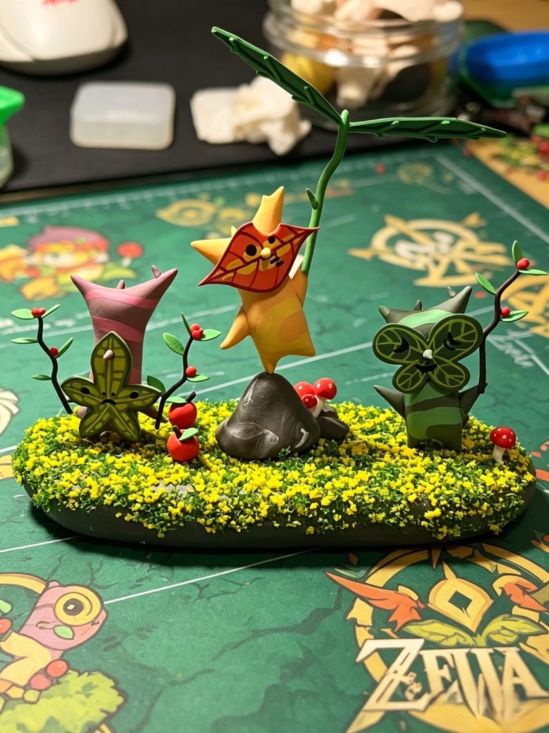 May include: A handcrafted diorama featuring three whimsical, stylized creatures amidst a miniature landscape. The scene includes a rock, small red mushrooms, and a base covered in yellow and green textured material. The creatures are in shades of green, orange, and pink.
