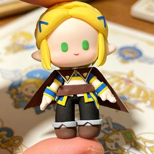 May include: A miniature figurine of a character with yellow hair, green eyes, and a small smile. The figure wears a brown cape, a blue and yellow tunic, black pants, and brown boots. The figure is held between two fingers.