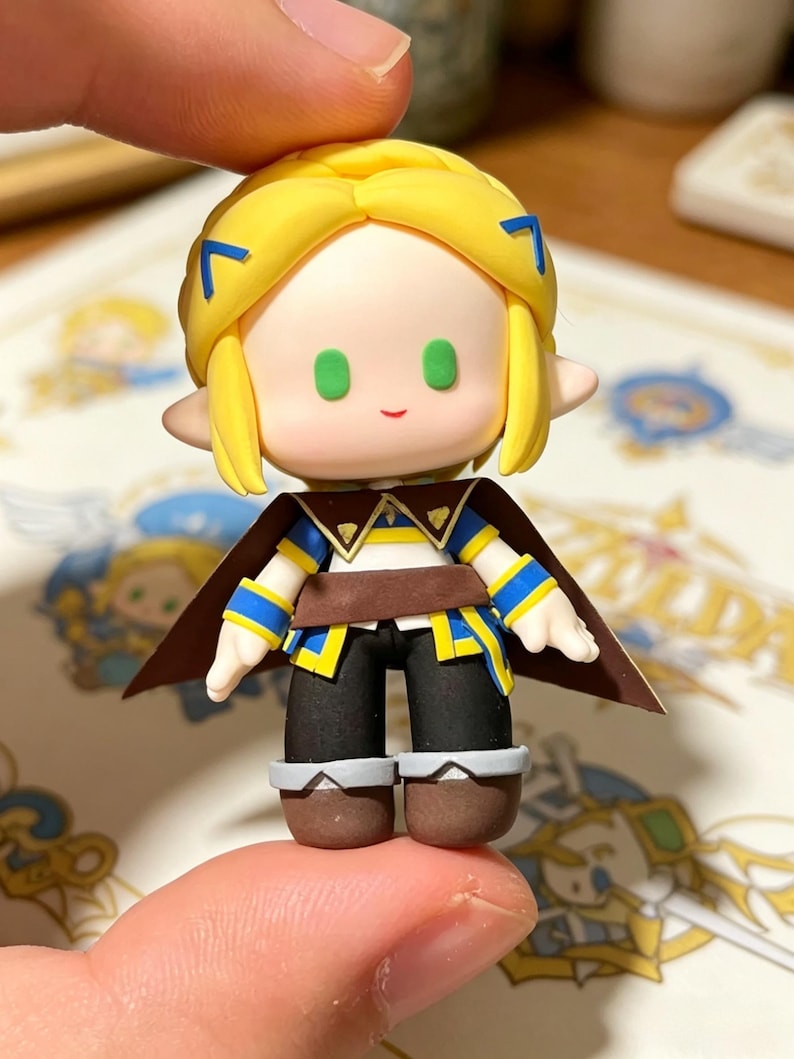 May include: A miniature figurine of a character with yellow hair, green eyes, and a small smile. The figure wears a brown cape, a blue and yellow tunic, black pants, and brown boots. The figure is held between two fingers.