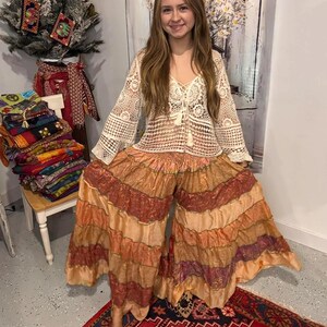 May include: A cream-colored crochet top with long sleeves and a V-neck, paired with wide-leg pants in shades of orange, brown, and gold. The pants have a layered, ruffled design. The outfit is bohemian-inspired.