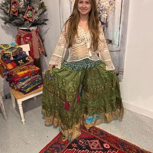 Può includere: Una donna indossa un top all'uncinetto color crema e pantaloni palazzo verdi fantasia. Una pila di tessuti colorati è su una piccola sedia bianca. Un albero di Natale è decorato con piccole tasche ricamate. Un tappeto rosso con un motivo dettagliato è sul pavimento.