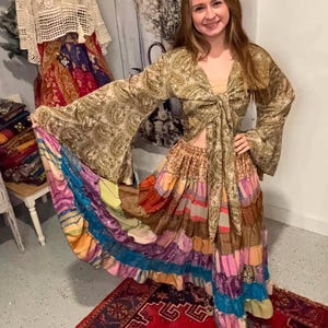May include: A long-sleeved, tie-front top in a gold and brown paisley print with wide bell sleeves, paired with a floor-length, tiered skirt featuring a patchwork of multicolored fabrics including stripes, floral patterns, and solid panels in various textures.