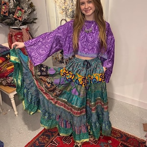 May include: A purple crop top with a swirling pattern and bell sleeves is paired with a tiered, patchwork skirt in shades of teal, purple, and gold. The skirt has a drawstring waist. The outfit is bohemian in style.