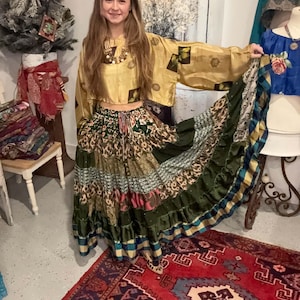 May include: A two-piece outfit consisting of a cropped, long-sleeved gold top with dark square accents and a long, tiered skirt featuring a patchwork of green, brown, and patterned fabrics with a drawstring waist and a ruffled hemline.