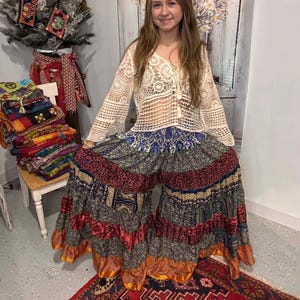 Può includere: Una donna indossa un top in crochet color crema con maniche lunghe e coulisse. Indossa pantaloni a gamba larga fantasia blu, rossi e oro, con balze. Un tappeto rosso fantasia è in primo piano. L'outfit è in stile bohémien.