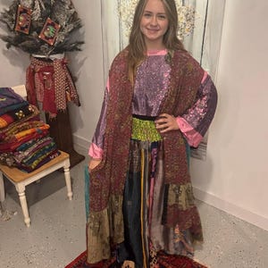 May include: A woman wearing a long, patchwork robe with a floral pattern in shades of burgundy, pink, and purple. The robe has wide sleeves and a contrasting pink trim. The dress underneath is a solid lavender color. The outfit is paired with a black belt with green and yellow accents.