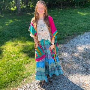 May include: A colorful bohemian outfit featuring a white crochet top, a long patchwork skirt in shades of blue, and a vibrant pink and turquoise kimono. The outfit is completed with patterned shoes, creating a unique and artistic look.