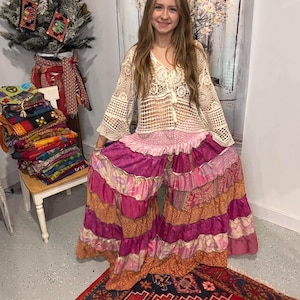 Puede incluir: Una mujer con un top de ganchillo color crema y pantalones de pernera ancha. Los pantalones son un patchwork de telas rosas, naranjas y moradas. Una pila de textiles coloridos está sobre una silla de madera.