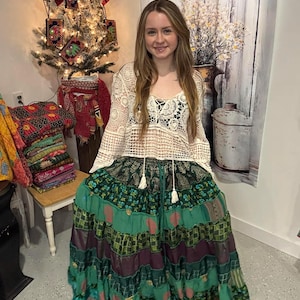 May include: A woman wearing a white crochet top over a black top and a long, tiered skirt. The skirt features panels of green, teal, and purple fabrics. The outfit is bohemian in style.