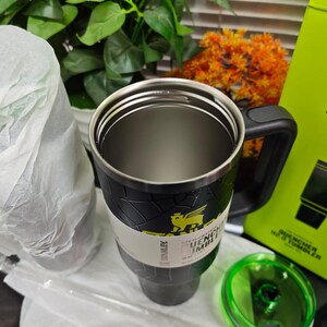 Pode incluir: Um copo Stanley Quencher H2.0 FlowState preto e prateado com uma al&ccedil;a preta e uma tampa verde. O copo tem um design texturizado e um logotipo amarelo. Uma caixa verde com o logotipo Stanley e um desenho do copo &eacute; vis&iacute;vel.