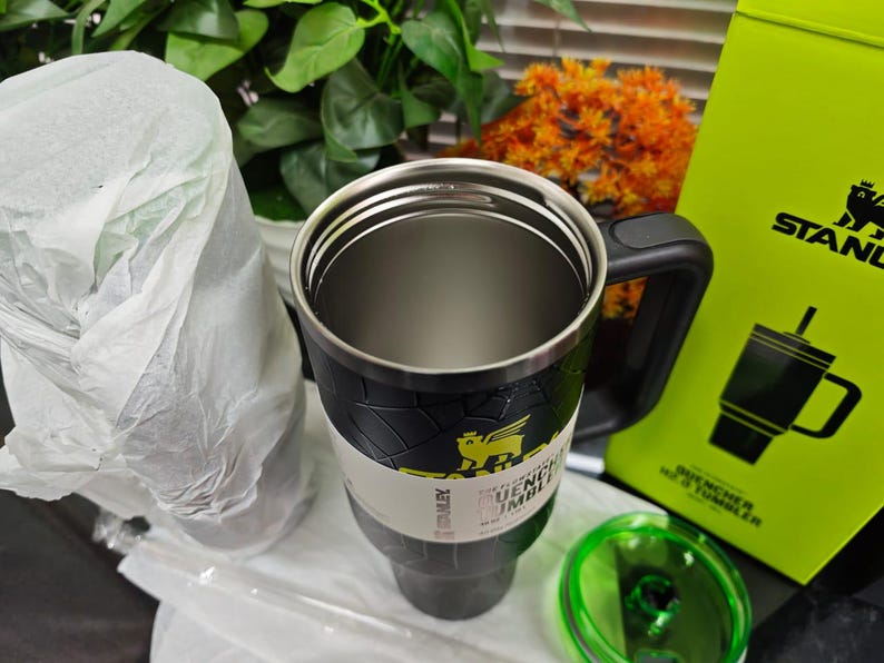 Pode incluir: Um copo Stanley Quencher H2.0 FlowState preto e prateado com uma al&ccedil;a preta e uma tampa verde. O copo tem um design texturizado e um logotipo amarelo. Uma caixa verde com o logotipo Stanley e um desenho do copo &eacute; vis&iacute;vel.