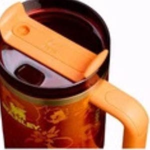May include: A stainless steel travel mug with an orange handle and lid. The mug has a floral design and the word "STANLEY" in gold lettering. The interior of the mug is dark brown.