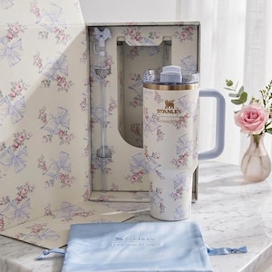 May include: A cream-colored Stanley tumbler with a floral and blue bow pattern, featuring a matching straw with a bow topper, a clear lid, and a light blue handle. The tumbler is presented in a matching floral box with a light blue drawstring pouch.