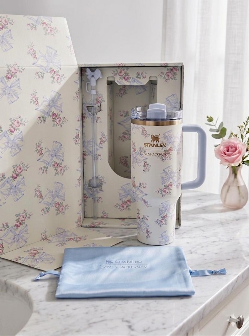 May include: A cream-colored Stanley tumbler with a floral and blue bow pattern, featuring a matching straw with a bow topper, a clear lid, and a light blue handle. The tumbler is presented in a matching floral box with a light blue drawstring pouch.