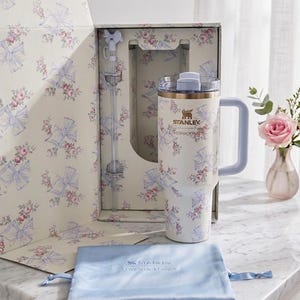 May include: A cream-colored Stanley tumbler with a floral and blue bow pattern, featuring a matching straw with a bow topper, a clear lid, and a light blue handle. The tumbler is presented in a matching floral box with a light blue drawstring pouch.