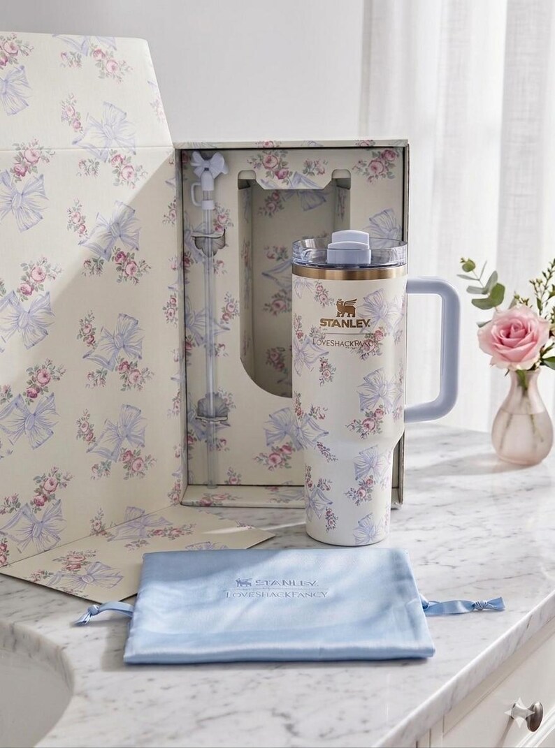 May include: A cream-colored Stanley tumbler with a floral and blue bow pattern, featuring a matching straw with a bow topper, a clear lid, and a light blue handle. The tumbler is presented in a matching floral box with a light blue drawstring pouch.